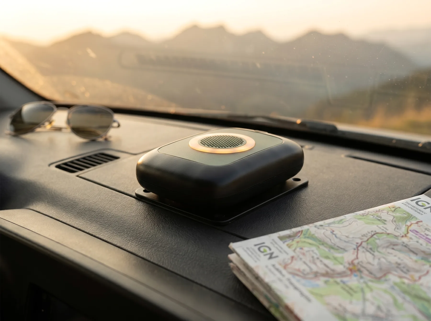 Le boîtier NomadGuard installé sur le tableau de bord d'un campervan, lunettes de soleil et carte IGN à proximité, montagnes au coucher de soleil